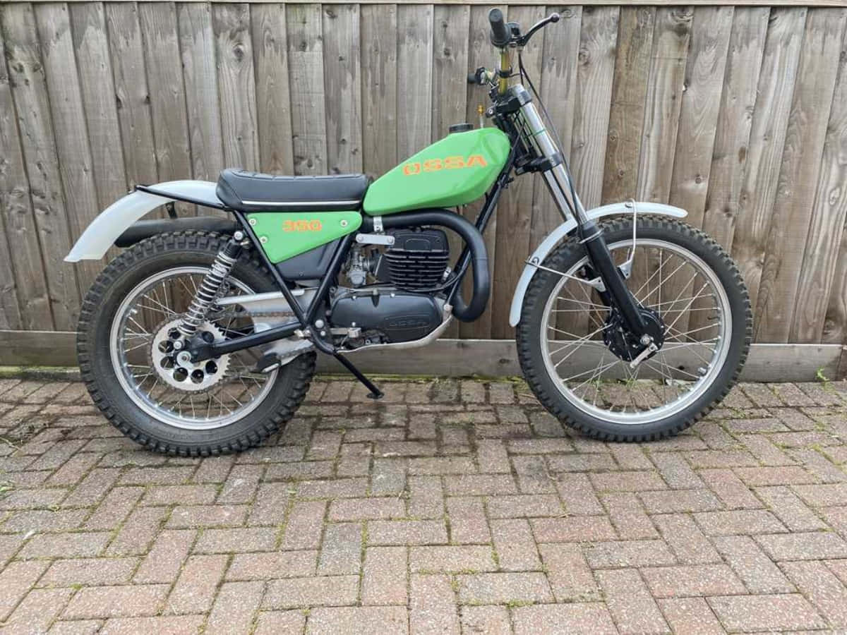 Ossa Motorcycle 桌布 壁紙 — 一輛復古綠色USAA 350cc越野機車停在磚砌庭院，背景是風化的木柵欄。