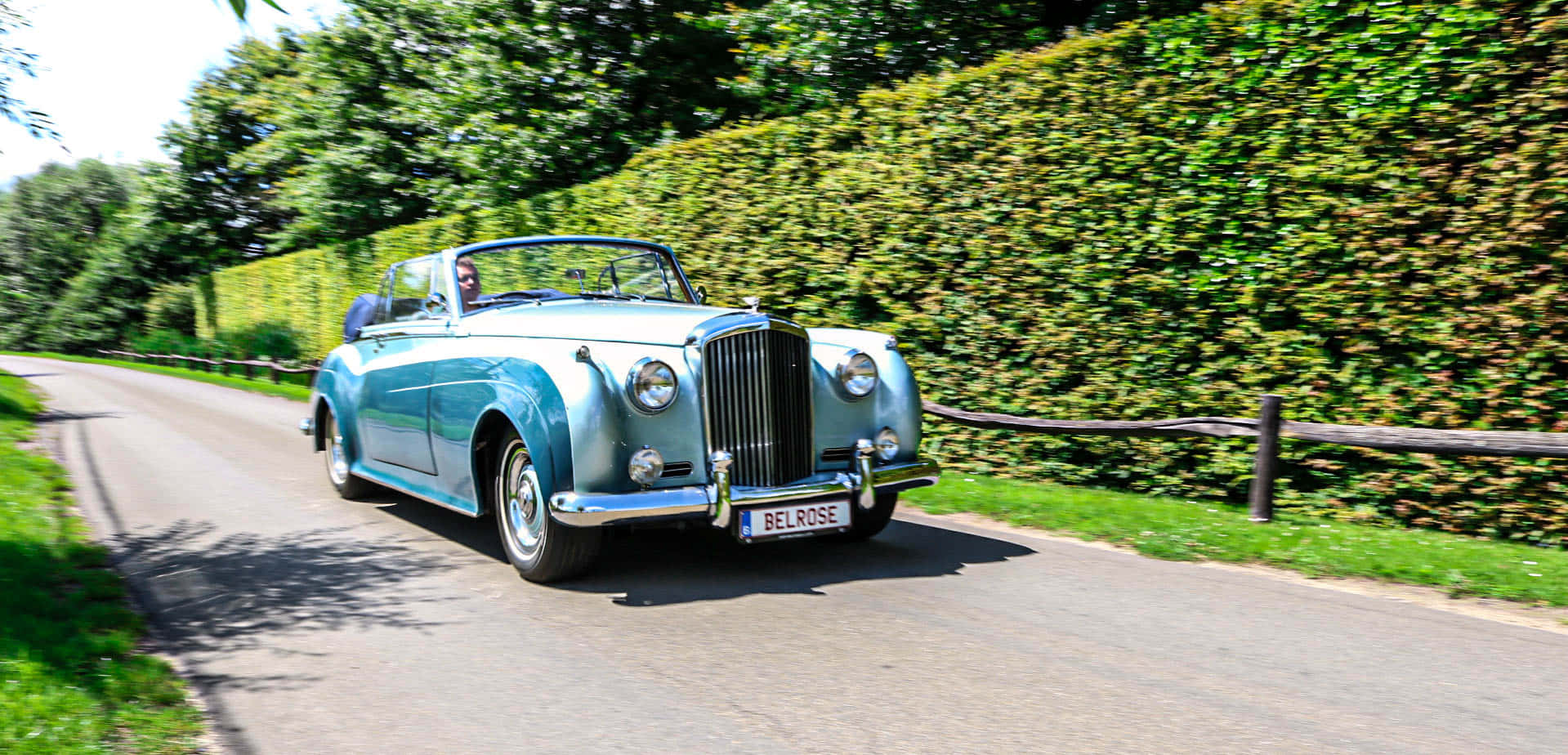 Bentley S2 桌布 壁紙 — 一輛復古淺藍色敞篷車在陽光明媚的鄉村道路上行駛，兩旁是茂密的綠色樹籬和樹木。
