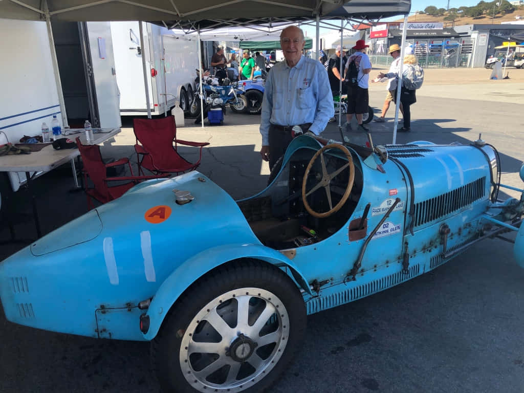 Bugatti Type 35 桌布 壁紙 — 一輛復古淺藍色賽車，引擎外露，木質方向盤，在陽光明媚的戶外車展的遮陽篷下，旁邊有一位微笑的男子。