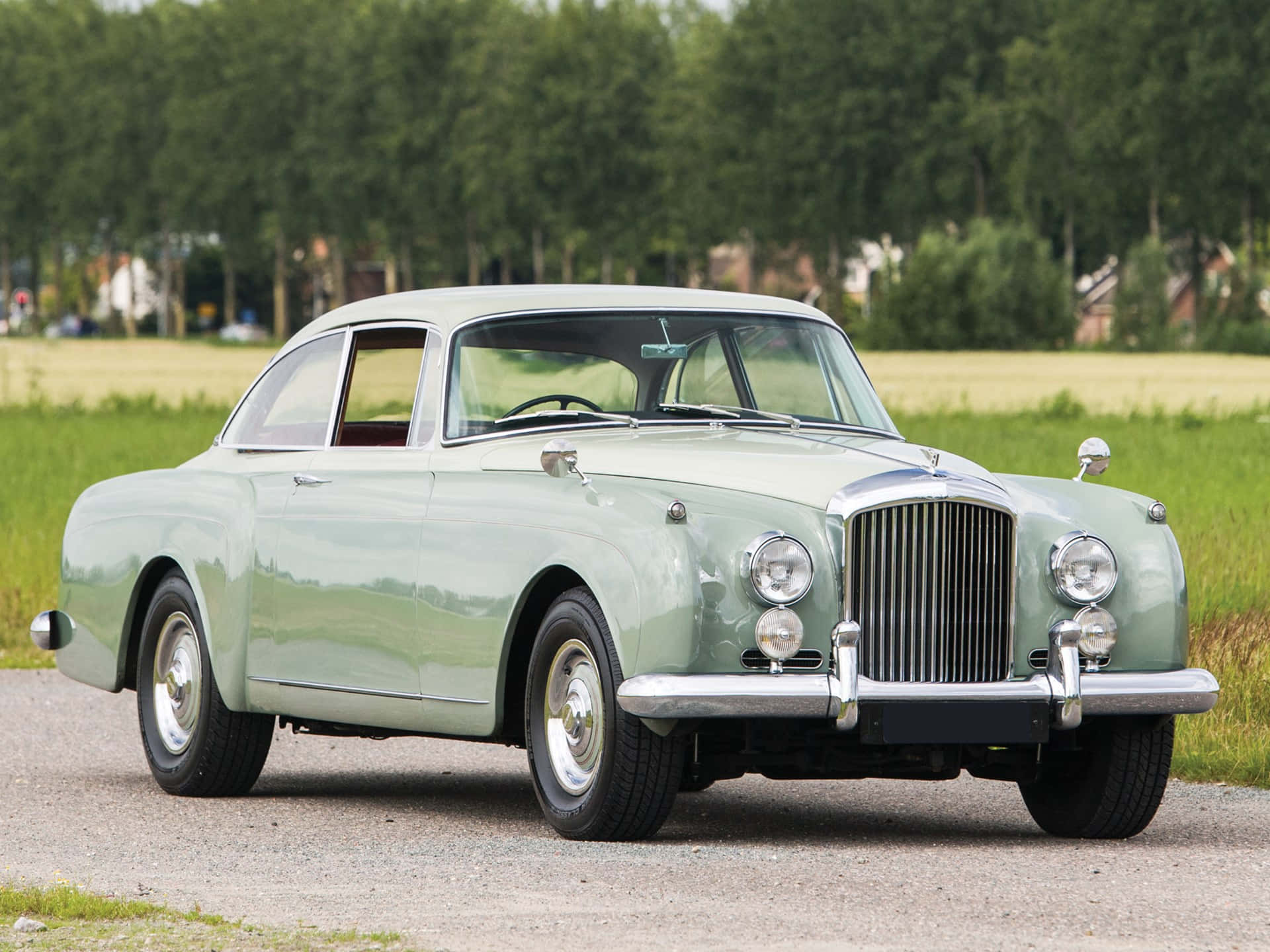 Bentley S2 桌布 壁紙 — 一輛復古薄荷綠色的賓利S1雙門轎跑車在日光下，在草地和樹木旁的碎石路上閃閃發光。
