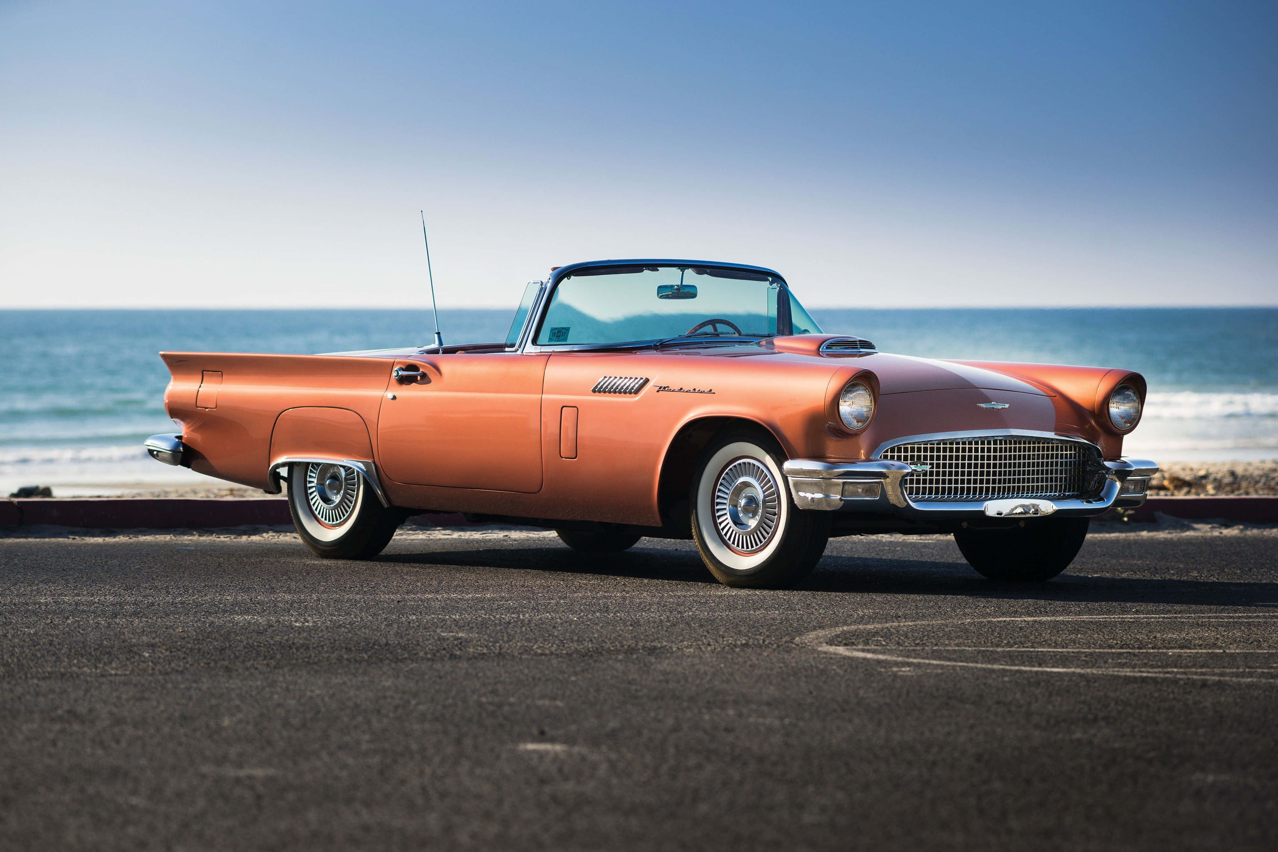 Ford Thunderbird 桌布 壁紙 — 一輛復古橙色敞篷車停泊在沿海公路上，背後有海浪拍打，天空清澈湛藍。