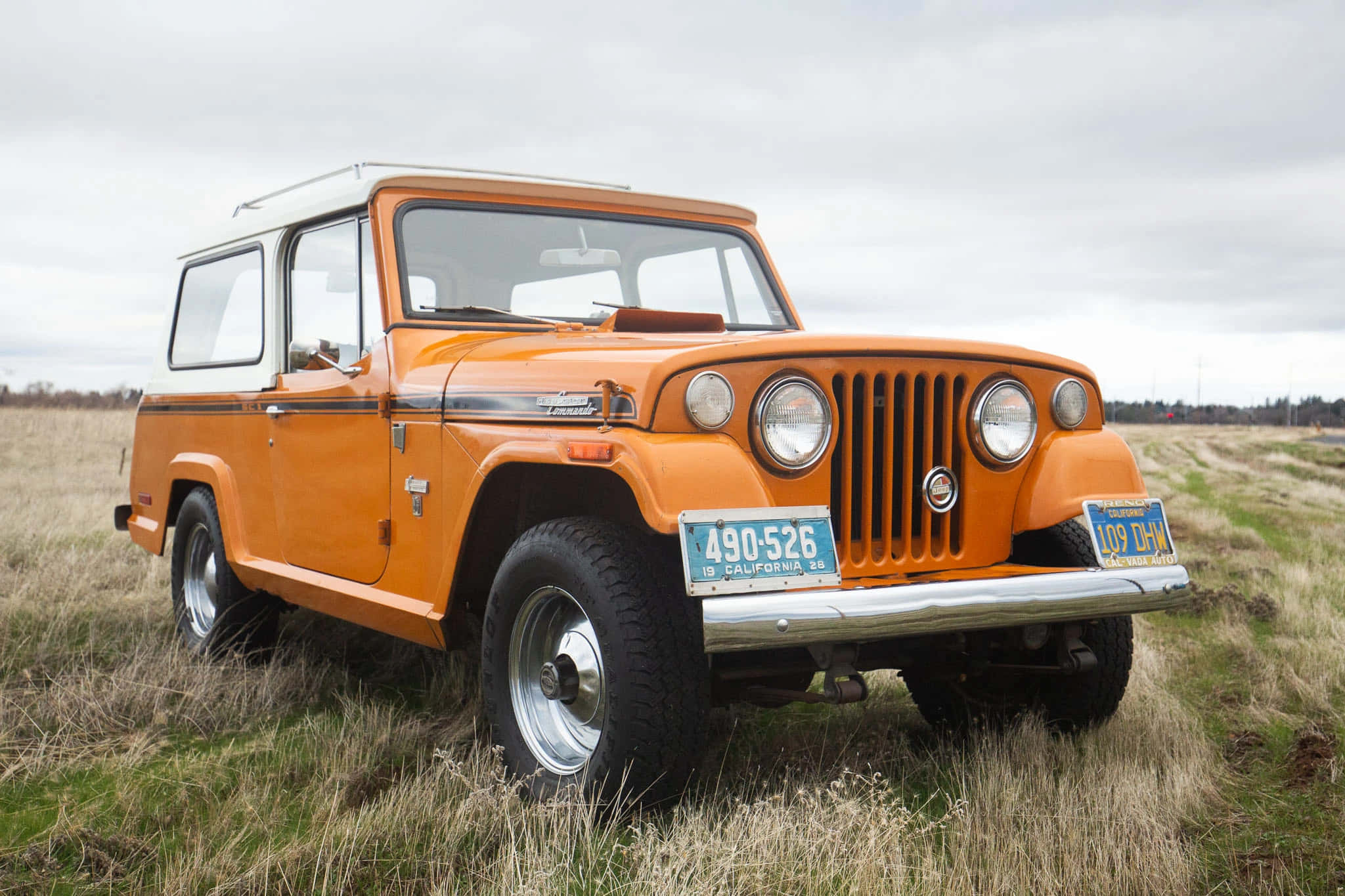Jeepster 桌布 壁紙 — 一輛復古橙色Jeep Wagoneer，白色車頂，停放在草地田野上，天空多雲。
