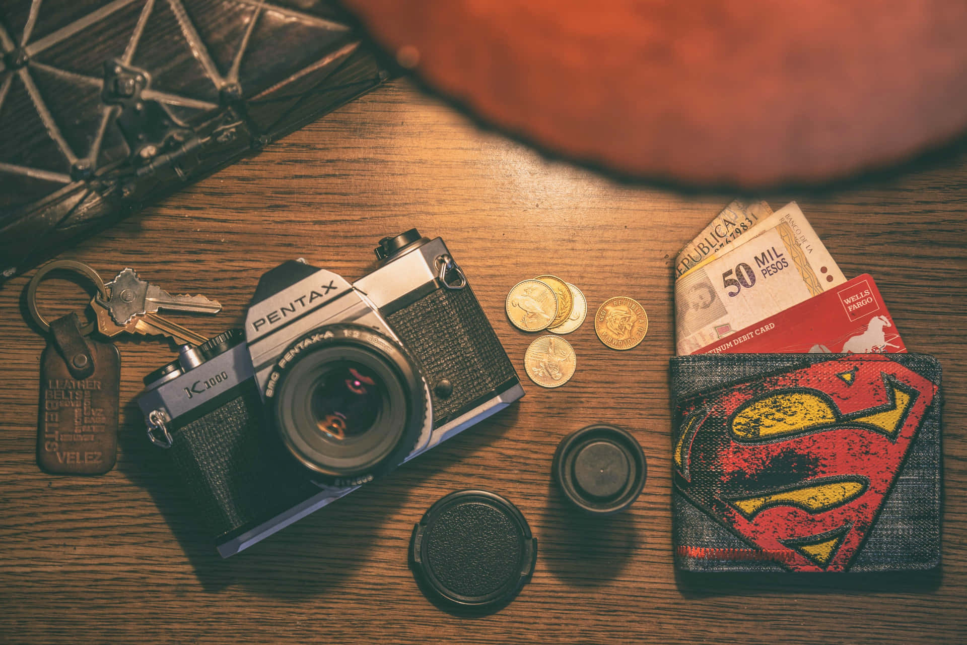  壁紙 — 一部復古的Pentax相機放在木桌上，旁邊有硬幣、鑰匙、超人錢包和貨幣，在溫暖的環境光下。