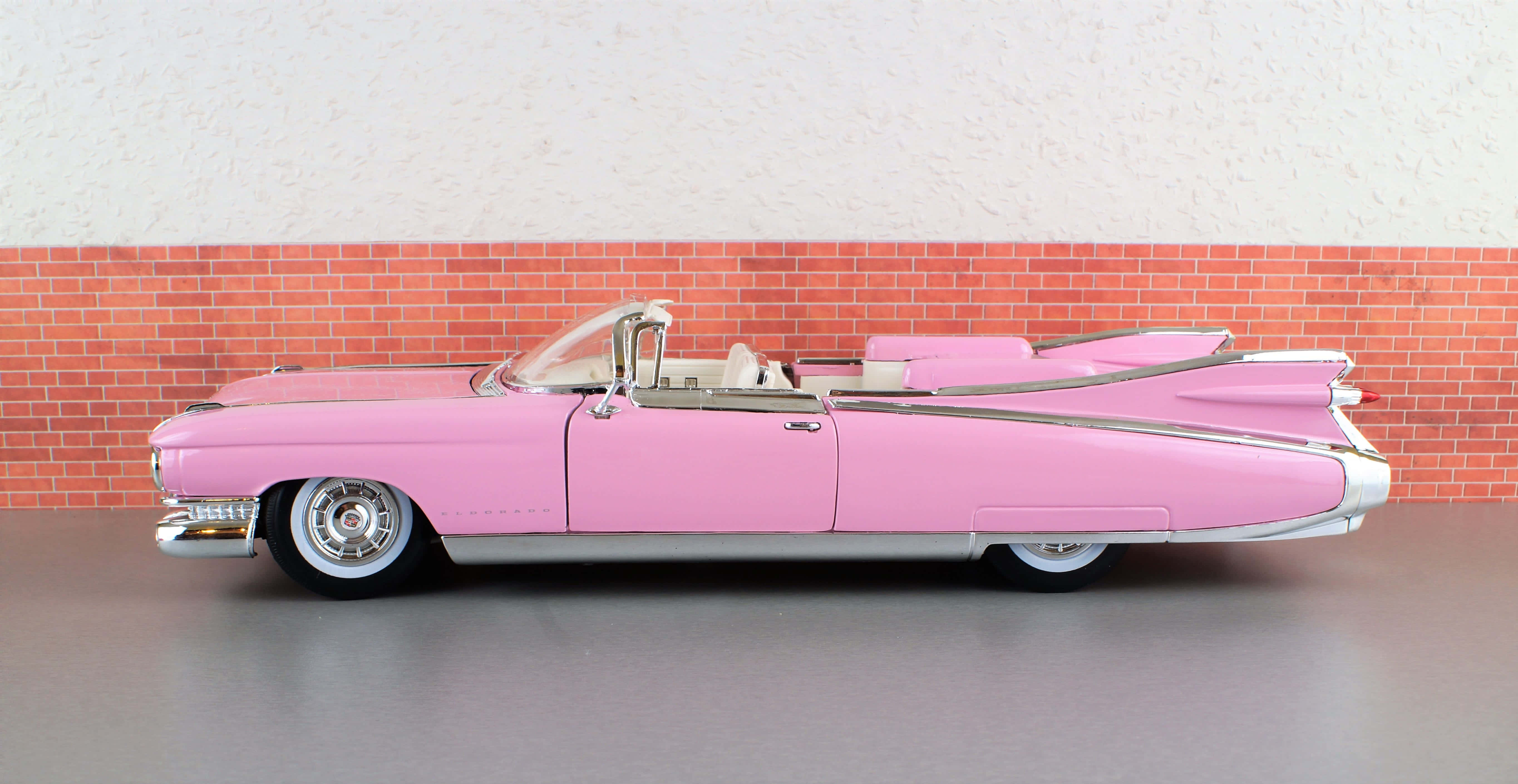 Cadillac Eldorado 桌布 壁紙 — 一輛帶有鍍鉻裝飾的復古粉紅色敞篷車優雅地停在紅磚牆和紋理白色上牆前。