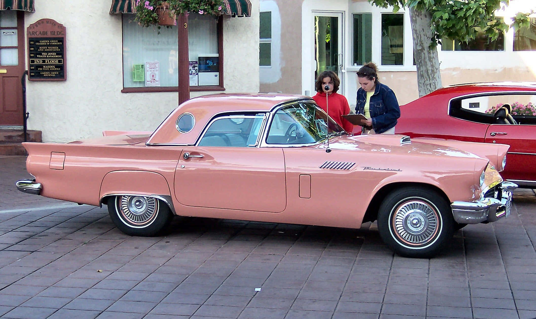 Ford Thunderbird 桌布 壁紙 — 一輛復古粉紅色 Thunderbird 經典汽車停在磚街上，兩名女性站在它旁邊，靠近一棟建築物。