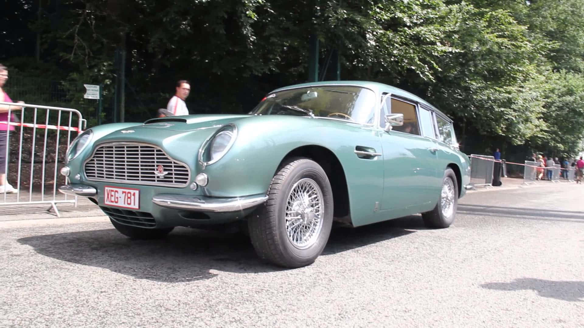 Aston Martin Db5 桌布 壁紙 — 一輛復古青綠色的奧斯頓馬丁DB5旅行車停在碎石路上，背景有樹木和觀眾。