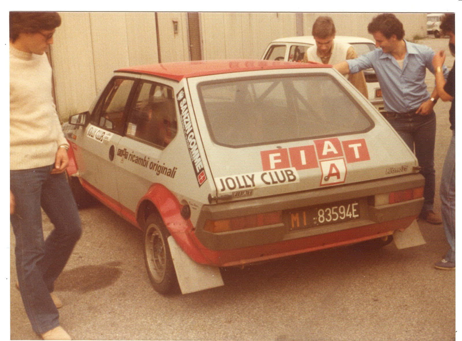 Fiat Ritmo 桌布 壁紙 — 一輛帶有 'Jolly Club' 貼紙的復古紅白菲亞特拉力賽車在車庫中被男士們包圍。