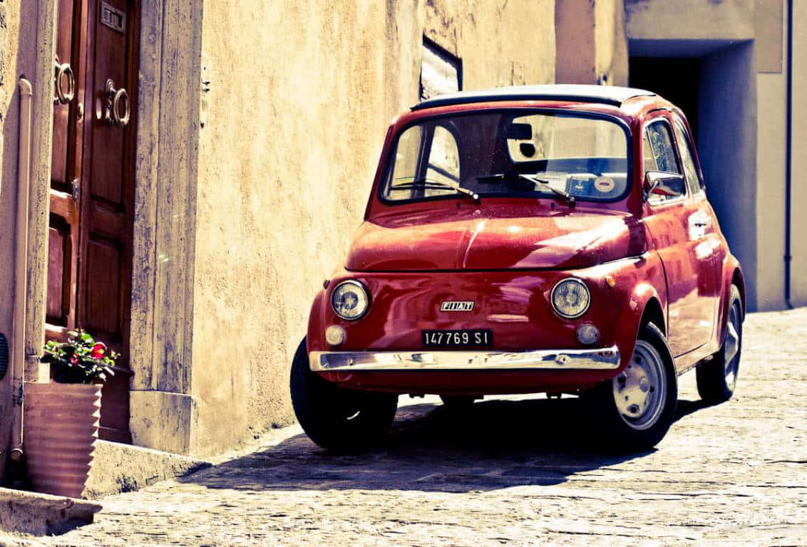 Fiat 500 桌布 壁紙 — 一輛復古紅色Fiat 500停放在陽光普照的鵝卵石街道上，旁邊是一棟有盆栽花卉的風化建築。