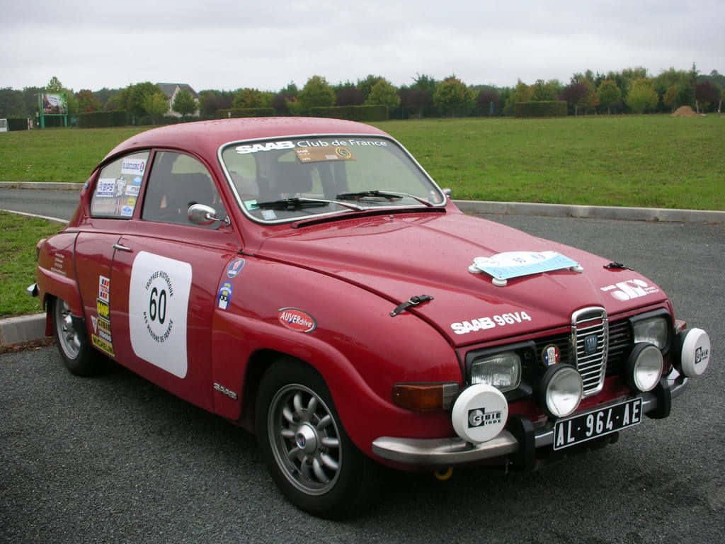 Saab 96 桌布 壁紙 — 一輛復古紅色薩博拉力賽車，編號60，停泊在瀝青路上，靠近草地，天空多雲。