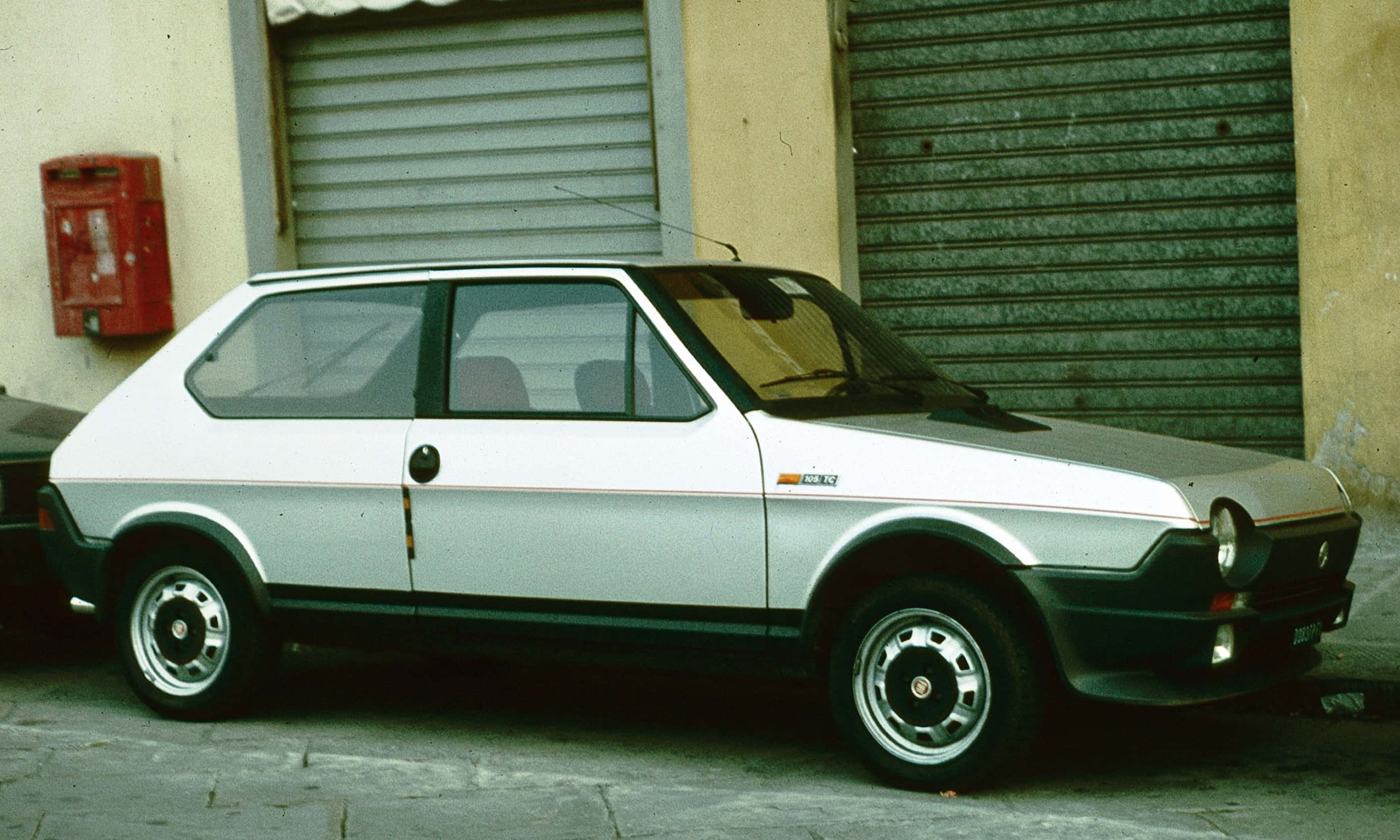 Fiat Ritmo 桌布 壁紙 — 一輛復古銀白色飛雅特 Panda 停泊在鵝卵石街道上，旁邊有一個紅色郵筒和金屬百葉窗。
