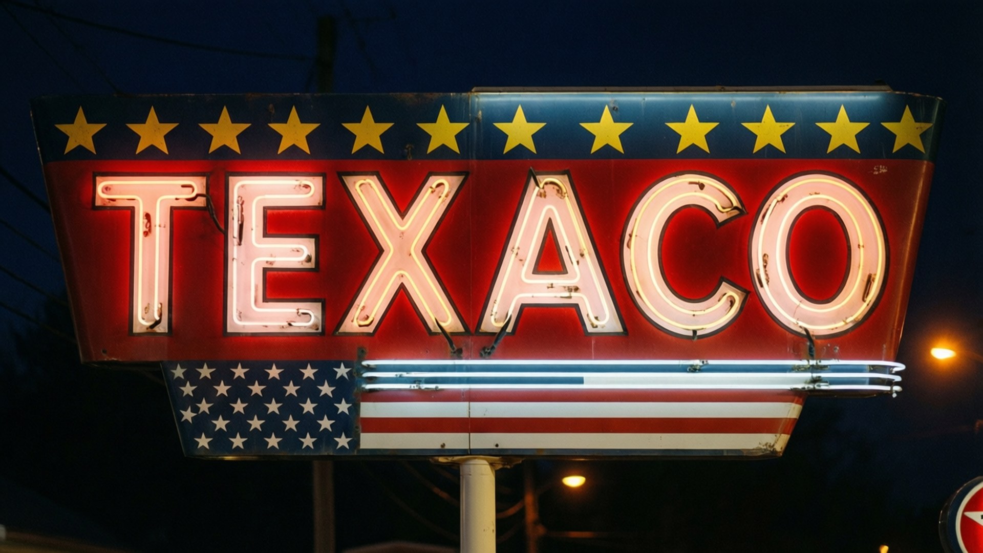 Texaco Neon Sign 壁紙 — 一個復古的Texaco霓虹燈牌在黑暗的夜空中發出紅、白、藍光，襯托著黃色星星和美國國旗圖案。