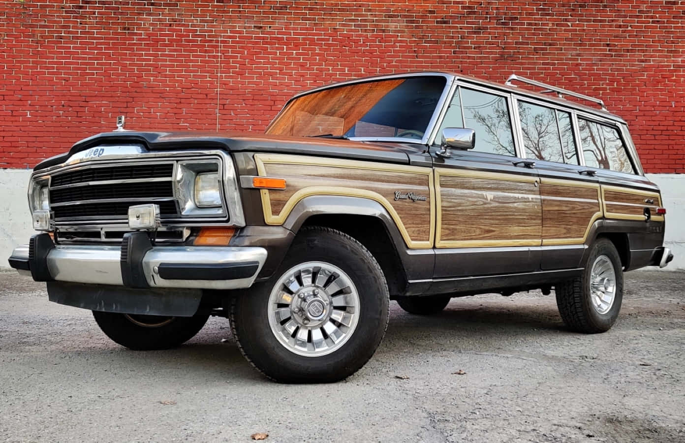Jeep Wagoneer 桌布 壁紙 — 一輛復古木紋車身的Jeep Grand Wagoneer停放在紅磚牆前的碎石地面上。