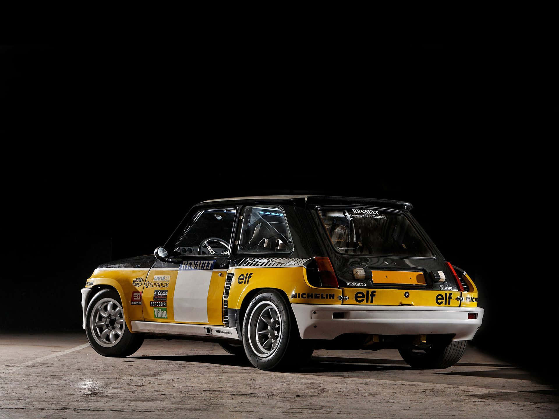 Renault 5 Turbo 桌布 壁紙 — 一輛復古黃黑配色的雷諾5 Turbo拉力賽車在黑暗的攝影棚背景下被戲劇性地照亮。