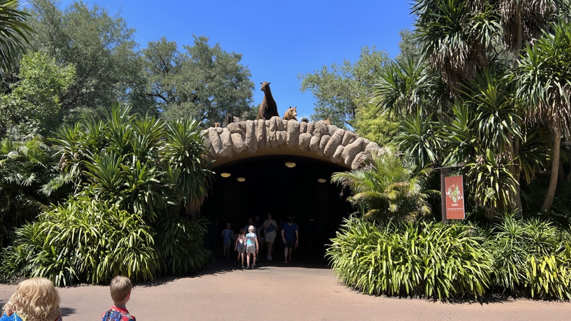 Houston Zoo Entrance 壁紙 — 訪客在裝飾著袋鼠雕像的石拱門下行走，周圍是茂密的綠色熱帶植物，頭頂是清澈的藍天。