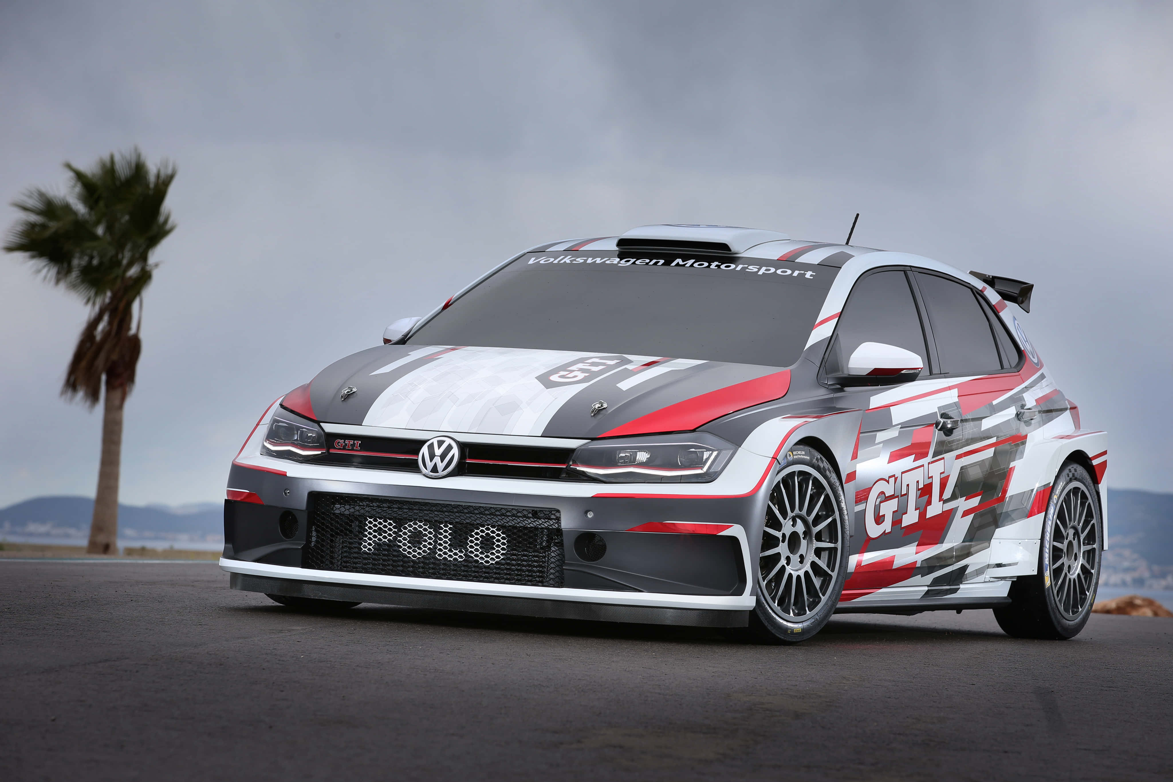 Volkswagen Polo 桌布 壁紙 — 一輛紅白黑塗裝的福斯Polo GTI拉力賽車停在柏油路上，旁邊有棕櫚樹，天空多雲。