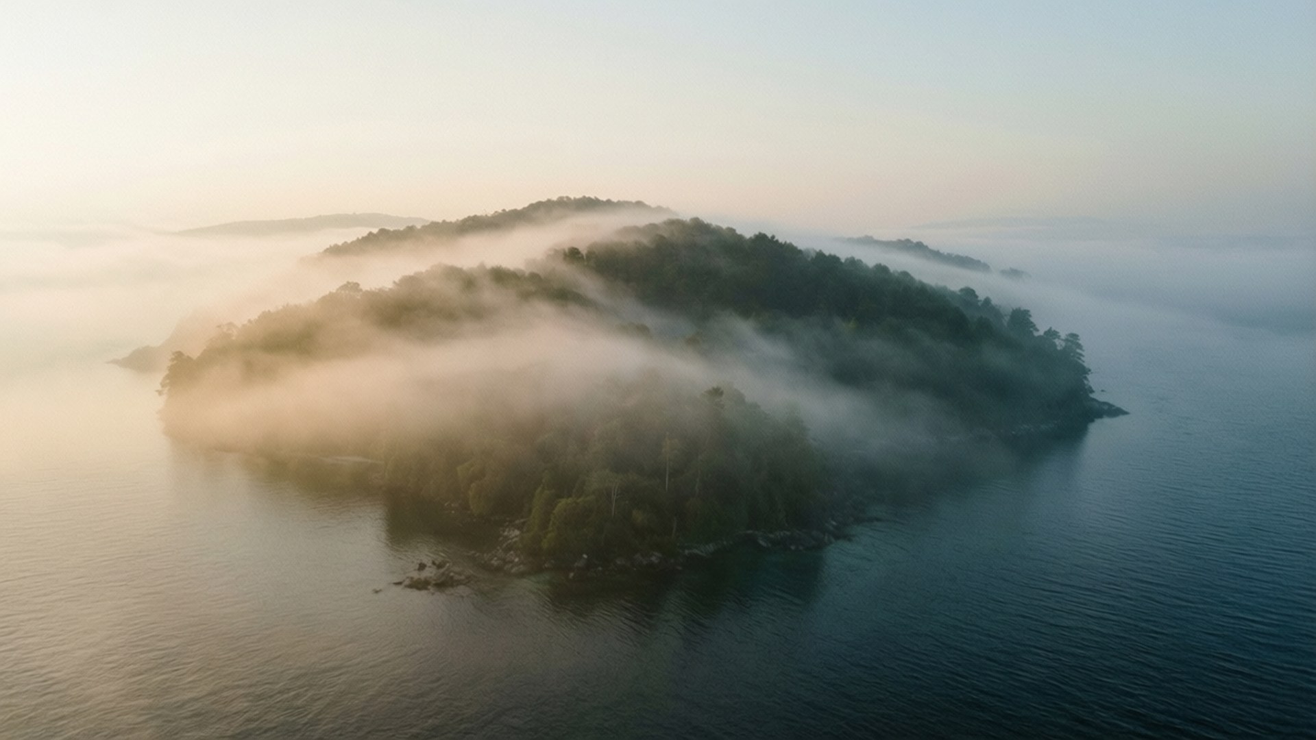 Misty Morning Island 壁紙 — 黎明時分，一座森林覆蓋的島嶼從平靜水域上的薄霧中浮現的鳥瞰圖。