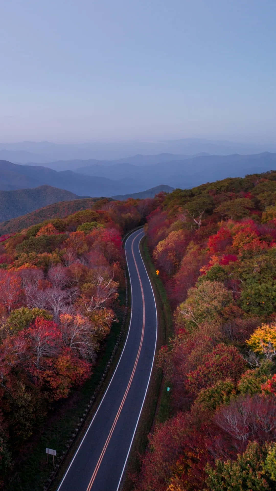  壁紙 — 一條蜿蜒的道路穿過色彩繽紛的秋日森林，層層疊疊的山巒逐漸消失在迷濛的藍色遠方。