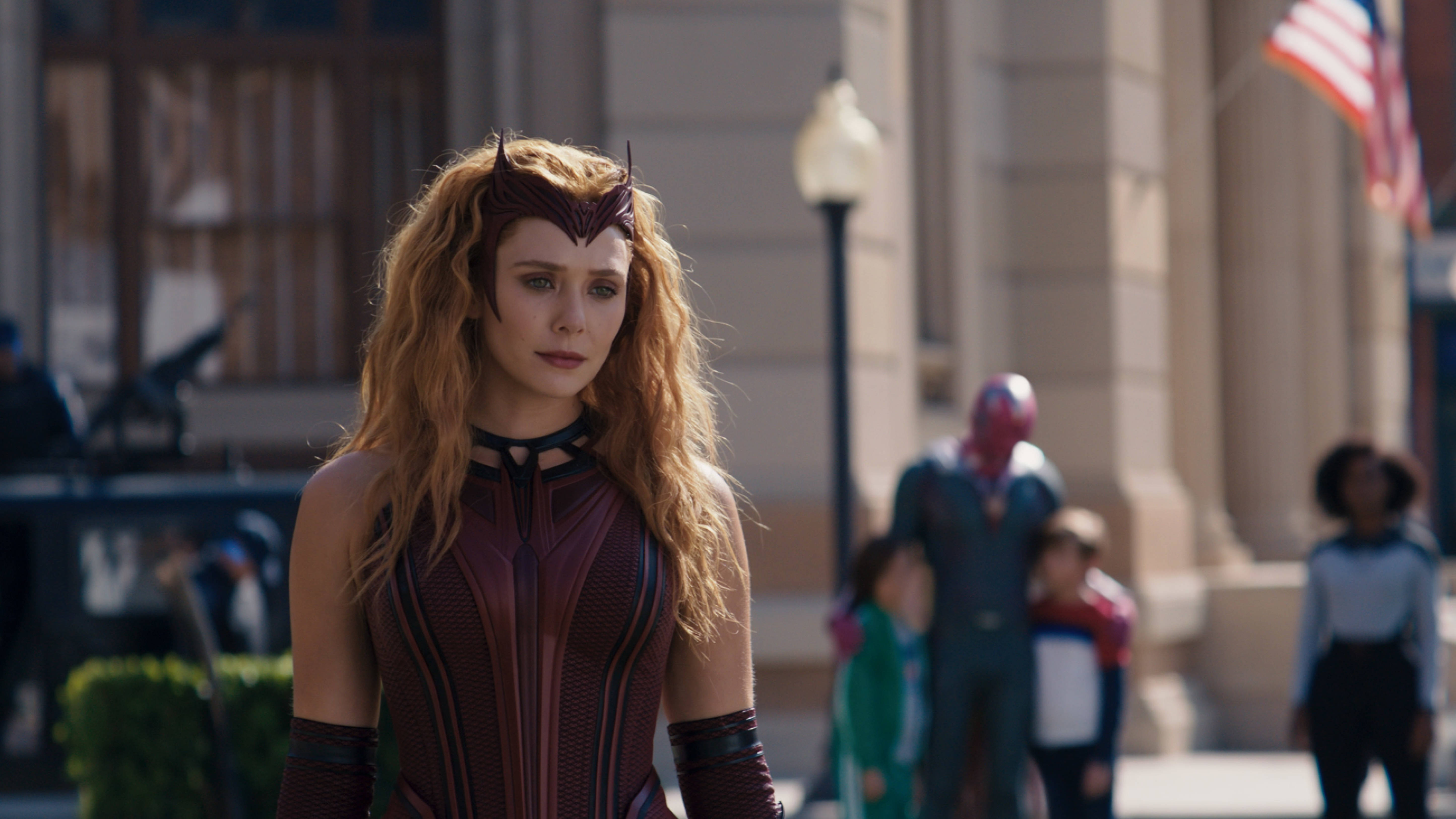 漫威 桌布 壁紙 — Wanda Maximoff 身穿紅黑戰衣，在陽光明媚的城市街道上清晰站立，背景有模糊的行人和美國國旗。
