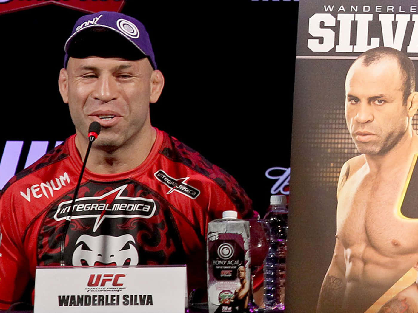 Champion 桌布 壁紙 — Wanderlei Silva 在UFC記者會上發言，身穿紅色品牌襯衫和紫色帽子，背景顯示他的名字。