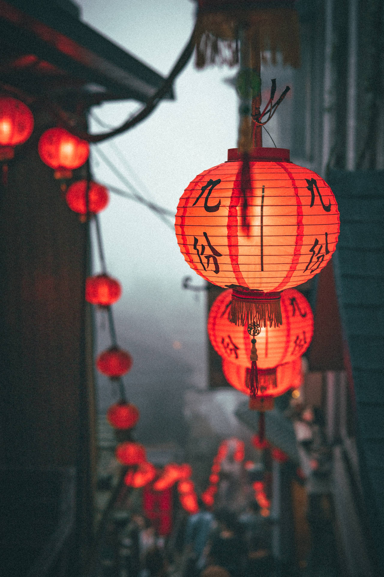 帶有黑色字符的紅色紙燈籠在黃昏時分霧氣瀰漫的小巷中溫暖地發光。 — 中文 壁紙