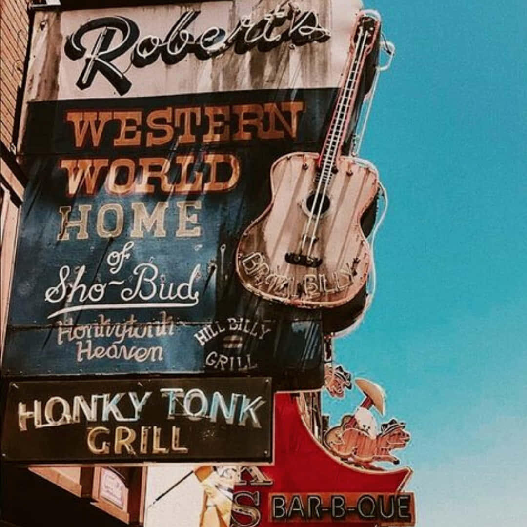 西部 桌布 壁紙 — Robert's Western World Honky Tonk Grill 的風化復古標誌，以吉他和牛仔圖案為特色，背景是明亮的藍天。