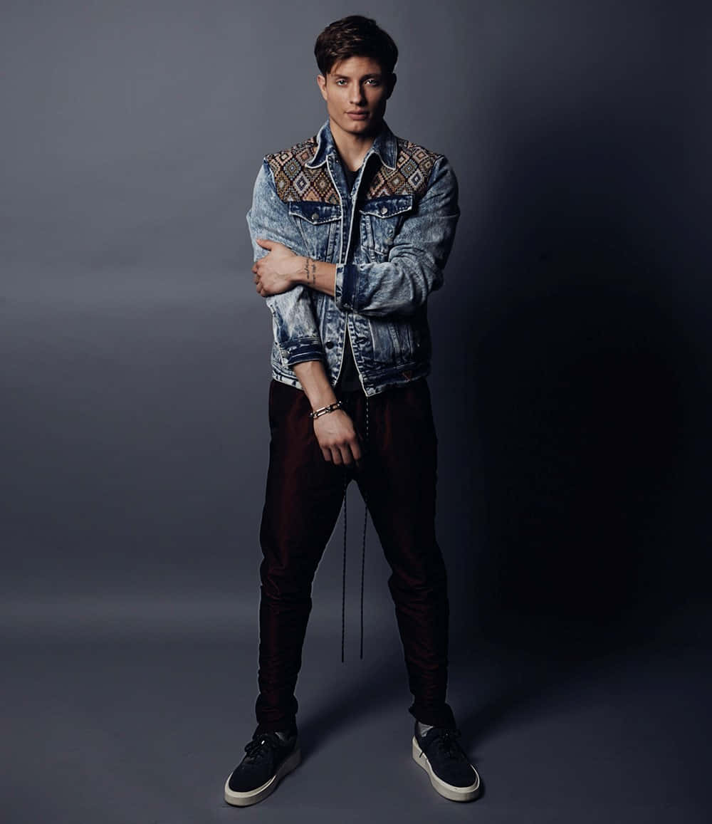 Matt Rife 桌布 壁紙 — 一位身穿圖案牛仔外套和深色褲子的年輕男子，在陰鬱的灰色工作室背景下自信地交叉雙臂站立。