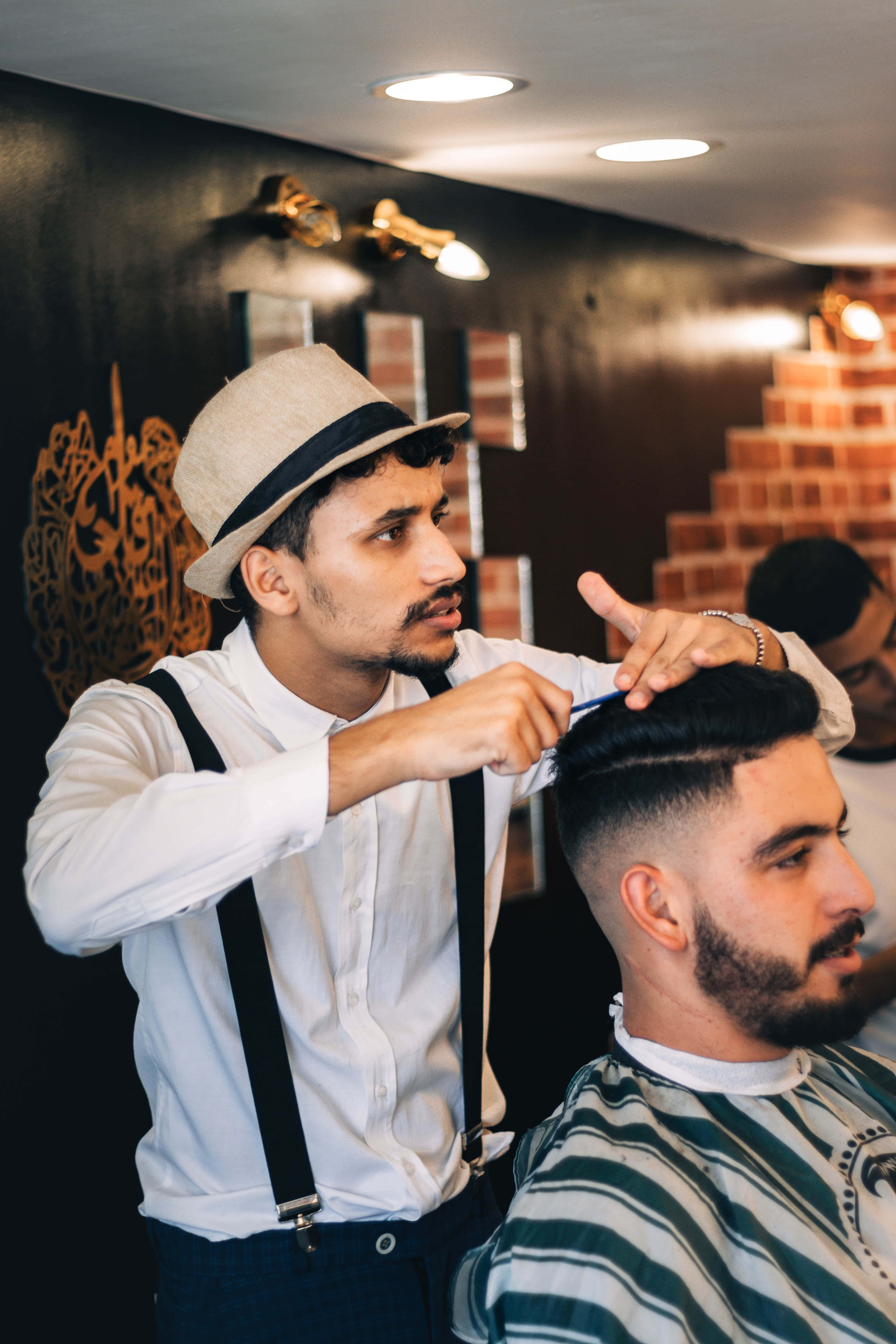 Barber 桌布 壁紙 — 一位戴著紳士帽和吊帶的理髮師，在溫暖燈光、磚牆裝飾的理髮店裡，細心地為客人修剪頭髮。