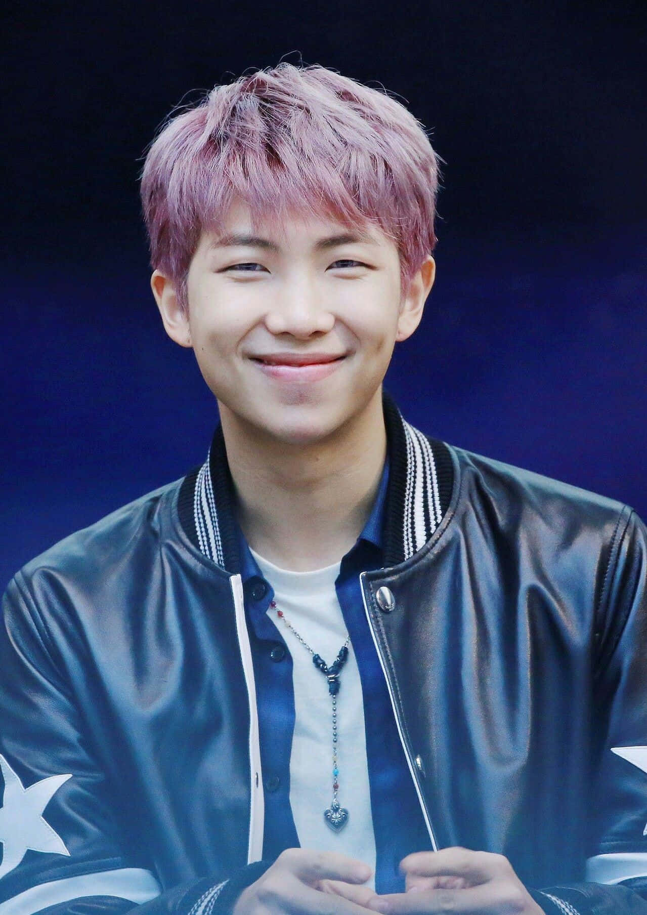 Bts Rap Monster 桌布 壁紙 — 一位粉紅色頭髮的年輕男子，穿著黑色皮夾克，在深藍色背景前溫暖地微笑。