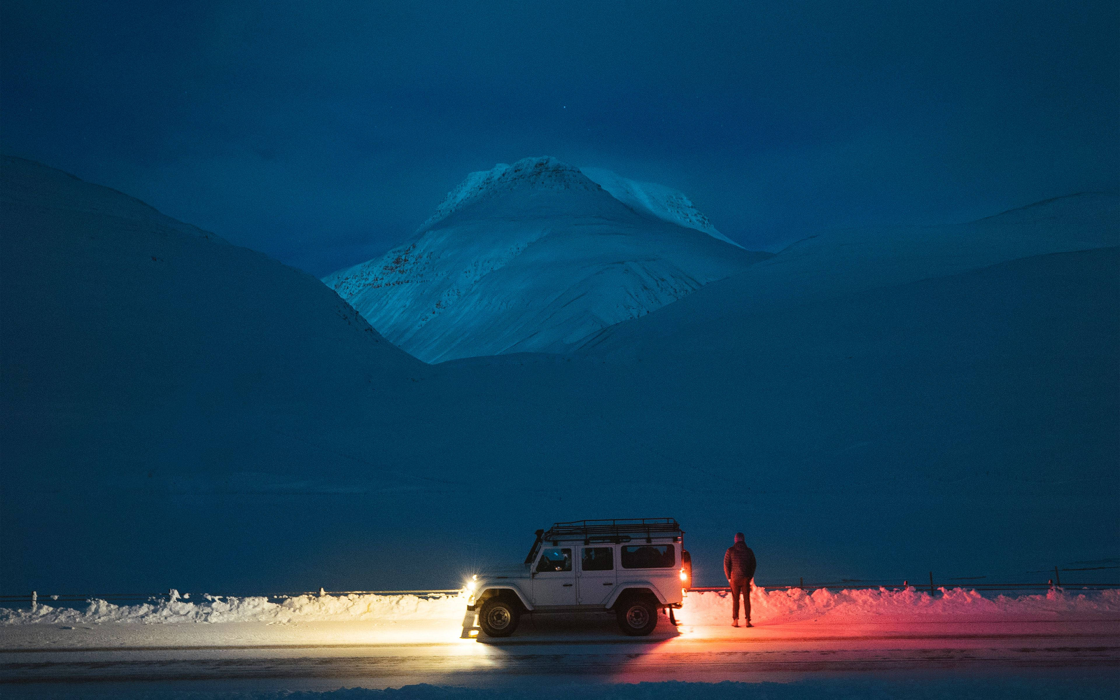 夜晚雪路上站在白色吉普車旁的人，背景有山。 — iPad 壁紙