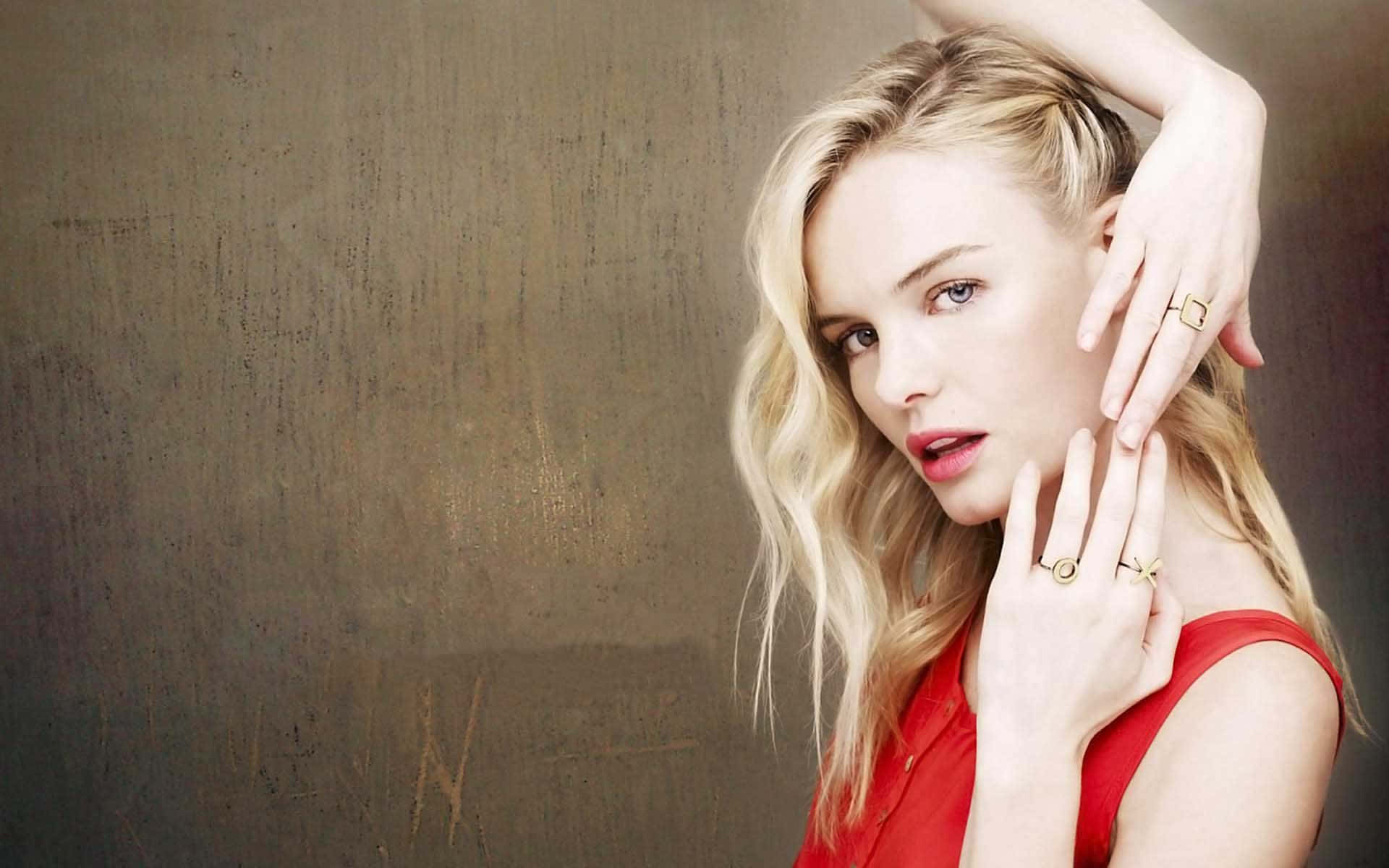 Kate Bosworth 桌布 壁紙 — 一位金髮女性身穿紅色上衣，在紋理豐富的棕色牆前擺姿勢，雙手靠近臉部，戴著戒指。