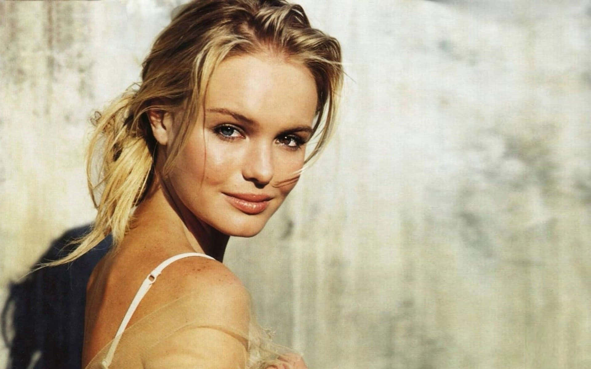 Kate Bosworth 桌布 壁紙 — 一位金髮女子帶著溫柔的微笑，在紋理混凝土牆前轉頭越過肩膀。