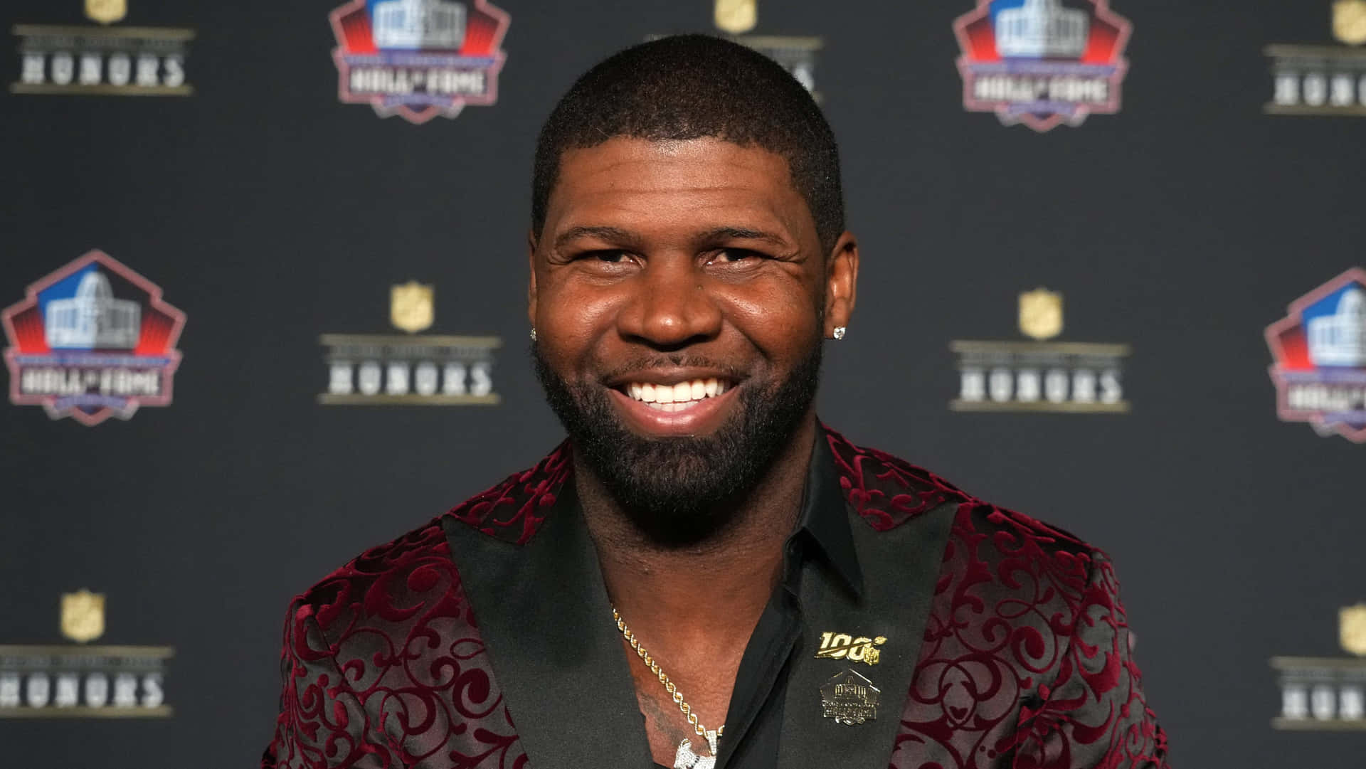  壁紙 — 一位留鬍鬚的微笑男士穿著紅色圖案外套，在NFL Honors活動中，背景為品牌佈景。