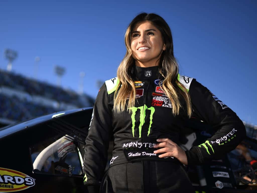  壁紙 — 一位面帶微笑的女性NASCAR車手，身穿黑色Monster Energy賽車服，在晴朗的藍天下自信地站在她的賽車旁。