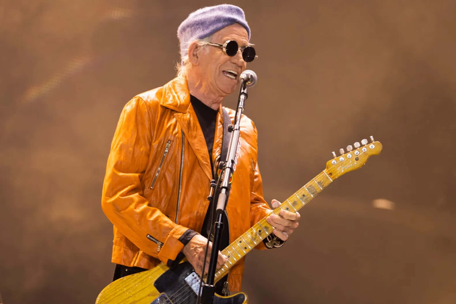  壁紙 — 一位年長男性穿著亮橙色皮夾克和紫色毛帽，在舞台上對著麥克風唱歌，同時彈奏黃色電吉他。