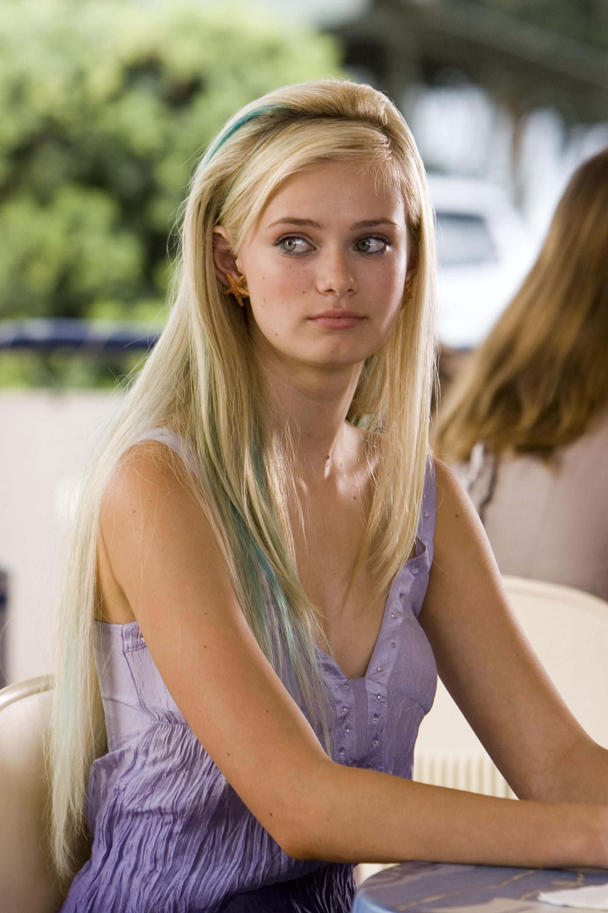 Sara Paxton 桌布 壁紙 — 一位年輕女性，長長的金髮帶有藍綠色挑染，穿著紫色連衣裙坐在桌旁，若有所思地望向別處。