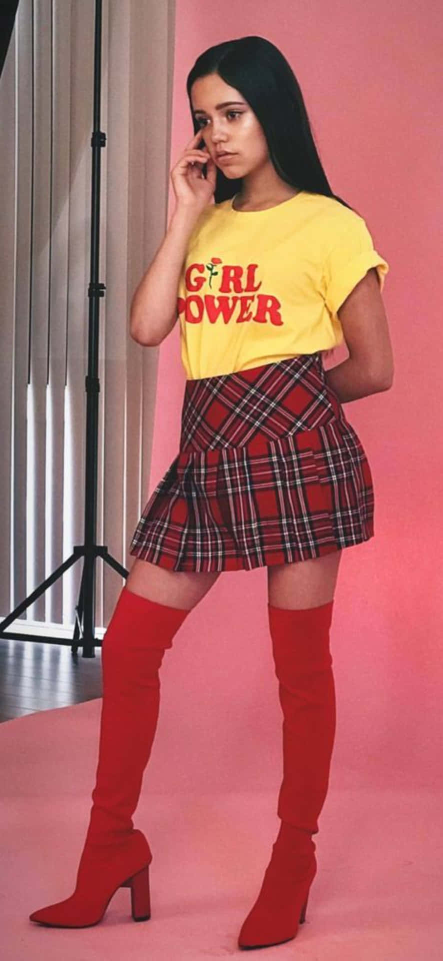  壁紙 — 一位年輕女性穿著黃色「girl power」T恤和紅色格紋裙，在粉紅色攝影棚背景前自信地擺姿勢。