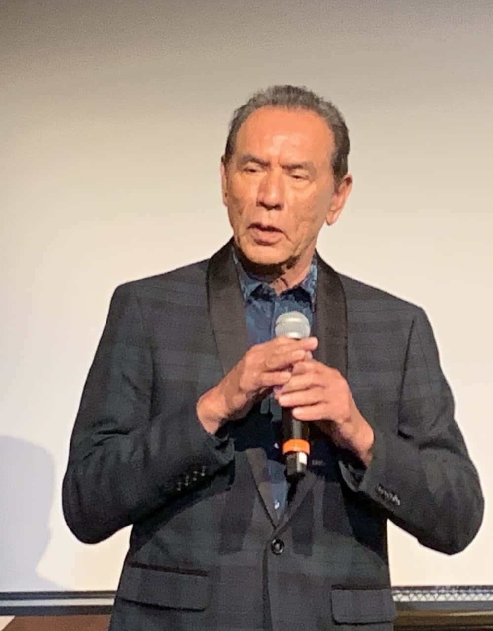  壁紙 — 一位年長男士身穿深色西裝，在純白色背景前對著麥克風說話。