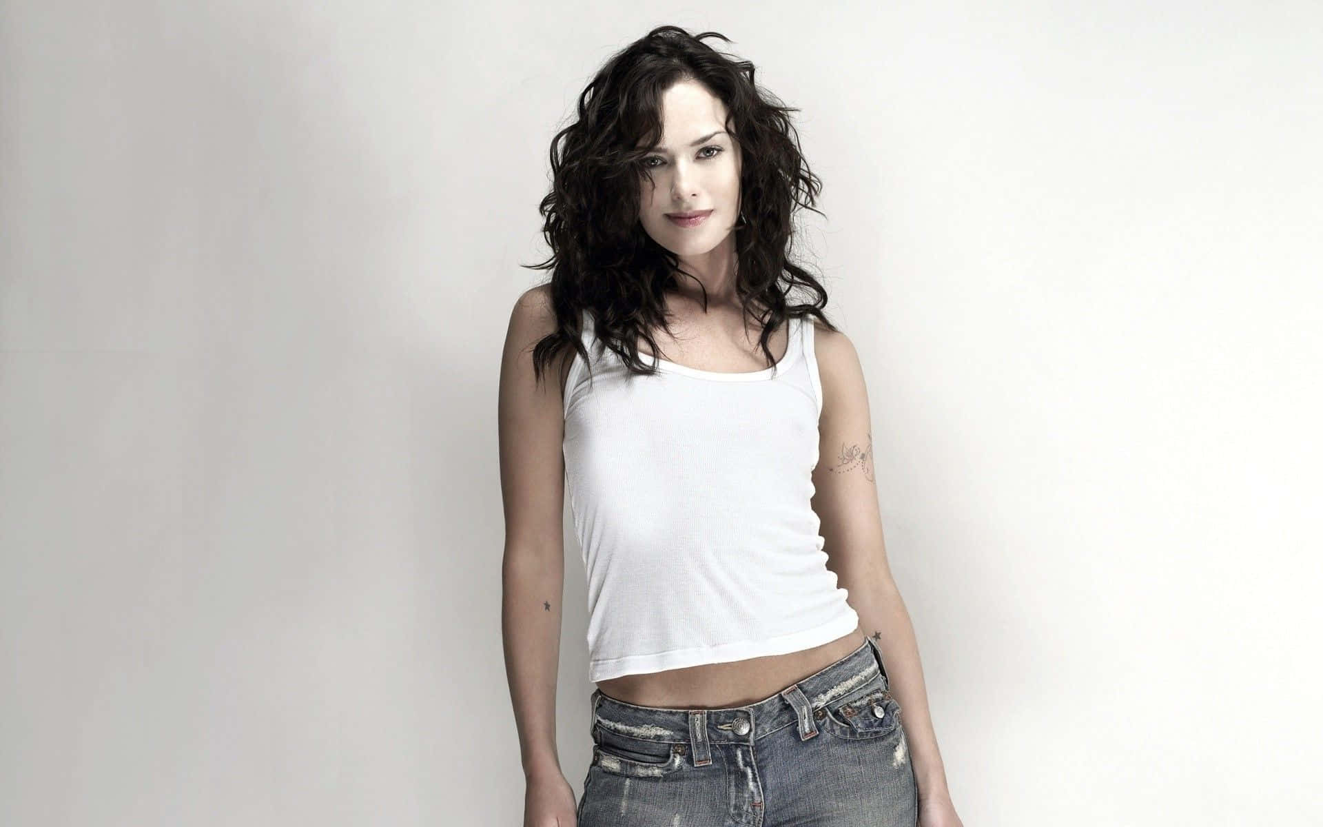 Lena Headey 桌布 壁紙 — 一位深色波浪長髮的女性穿著白色背心和破洞牛仔褲，站在純白背景前。