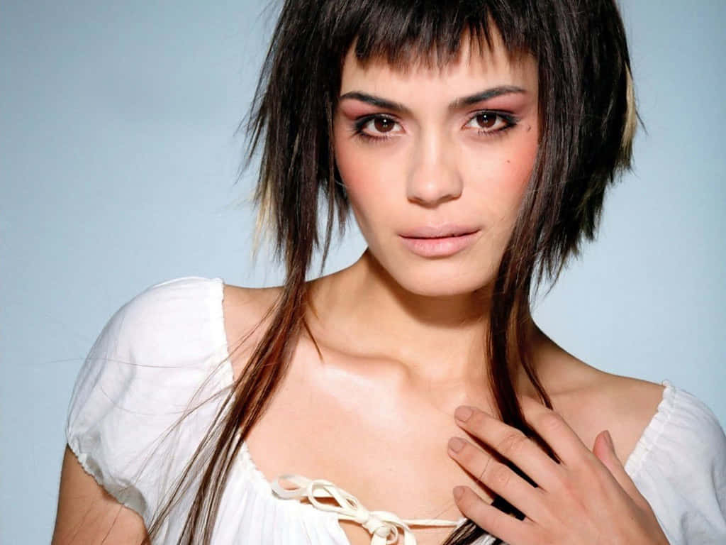 Shannyn Sossamon 桌布 壁紙 — 一位深色凌亂頭髮、妝容大膽的女性，穿著白色露肩上衣，在柔和的藍色背景前直視鏡頭。
