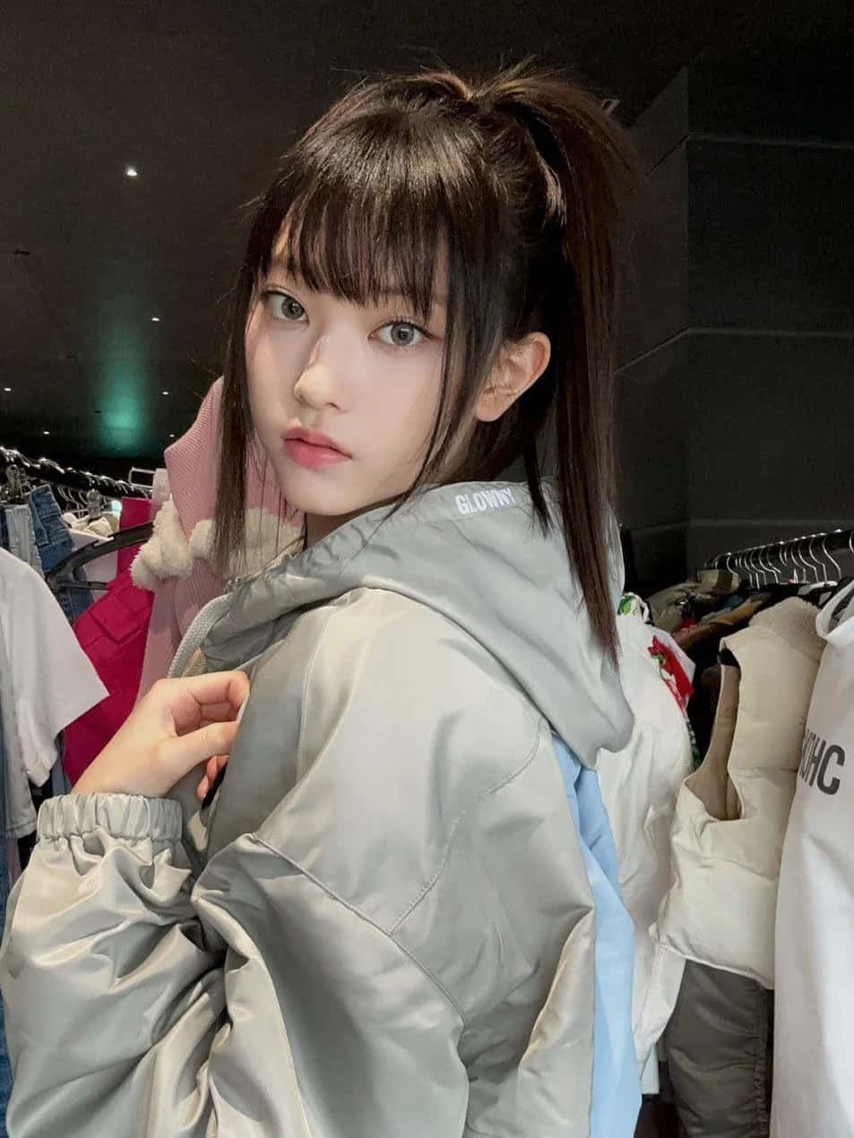  壁紙 — 一位深色頭髮高馬尾的年輕女性，在燈光昏暗的服裝店裡穿著淺灰色外套，回頭凝視。