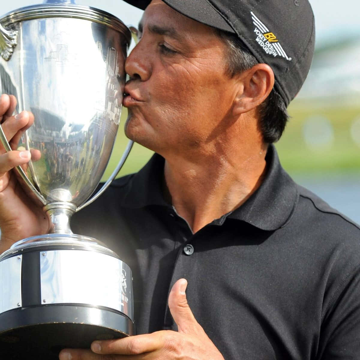 壁紙 — 一位勝利的哥爾夫球手在明亮的日光下，戴著黑色帽子和Polo衫，親吻閃閃發光的銀色獎盃。