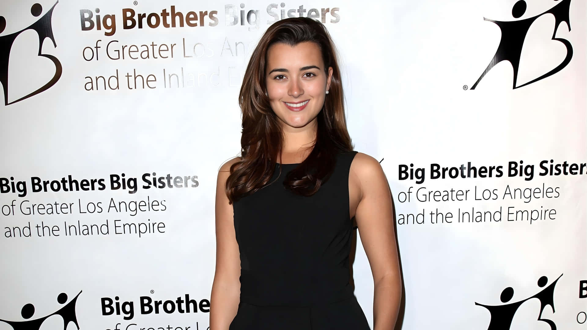  壁紙 — 一位微笑的黑裙女士自信地站在Big Brothers Big Sisters活動中，背景帶有品牌標誌。