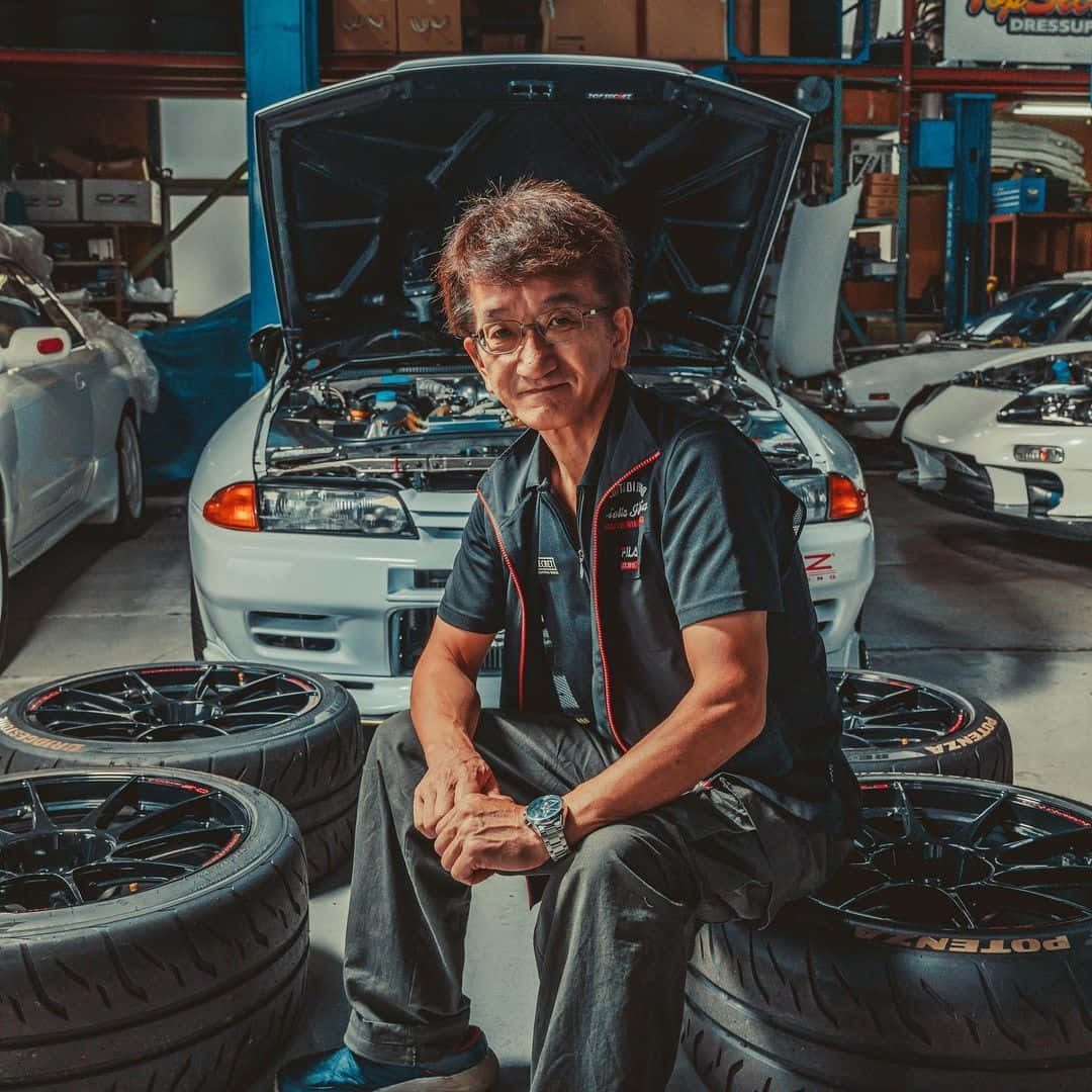  壁紙 — 一位微笑的機械師坐在輪胎上，車庫裡環繞著改裝車和打開的引擎蓋。