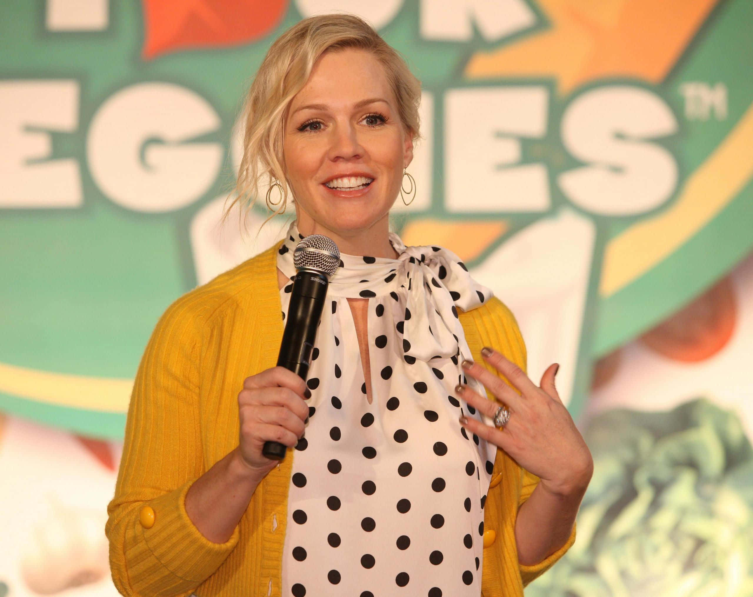 Jennie Garth 桌布 壁紙 — 一位微笑的金髮女性穿著黃色開襟毛衣和圓點襯衫，在Veggie Tales活動中拿著麥克風。