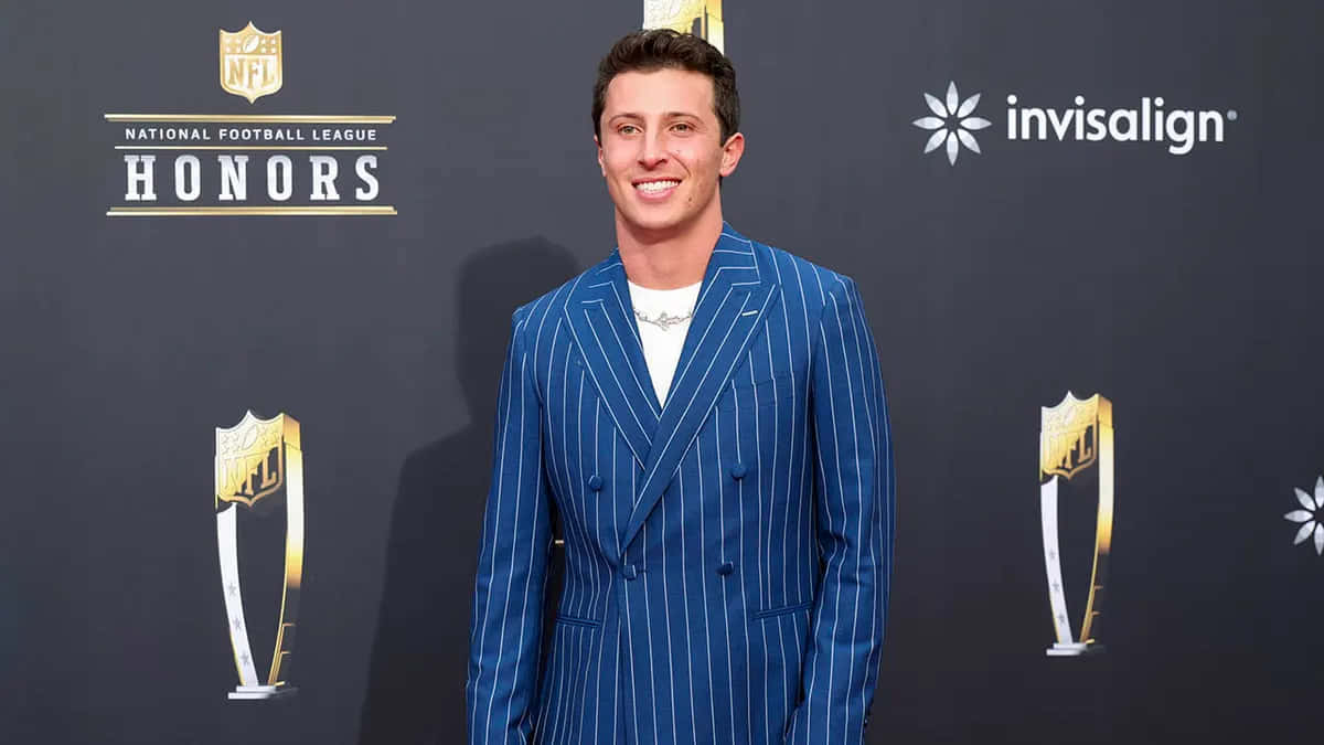  壁紙 — 一位微笑的男子身穿藍色細條紋西裝，自信地站在NFL Honors活動現場，身後有獎盃和標誌。