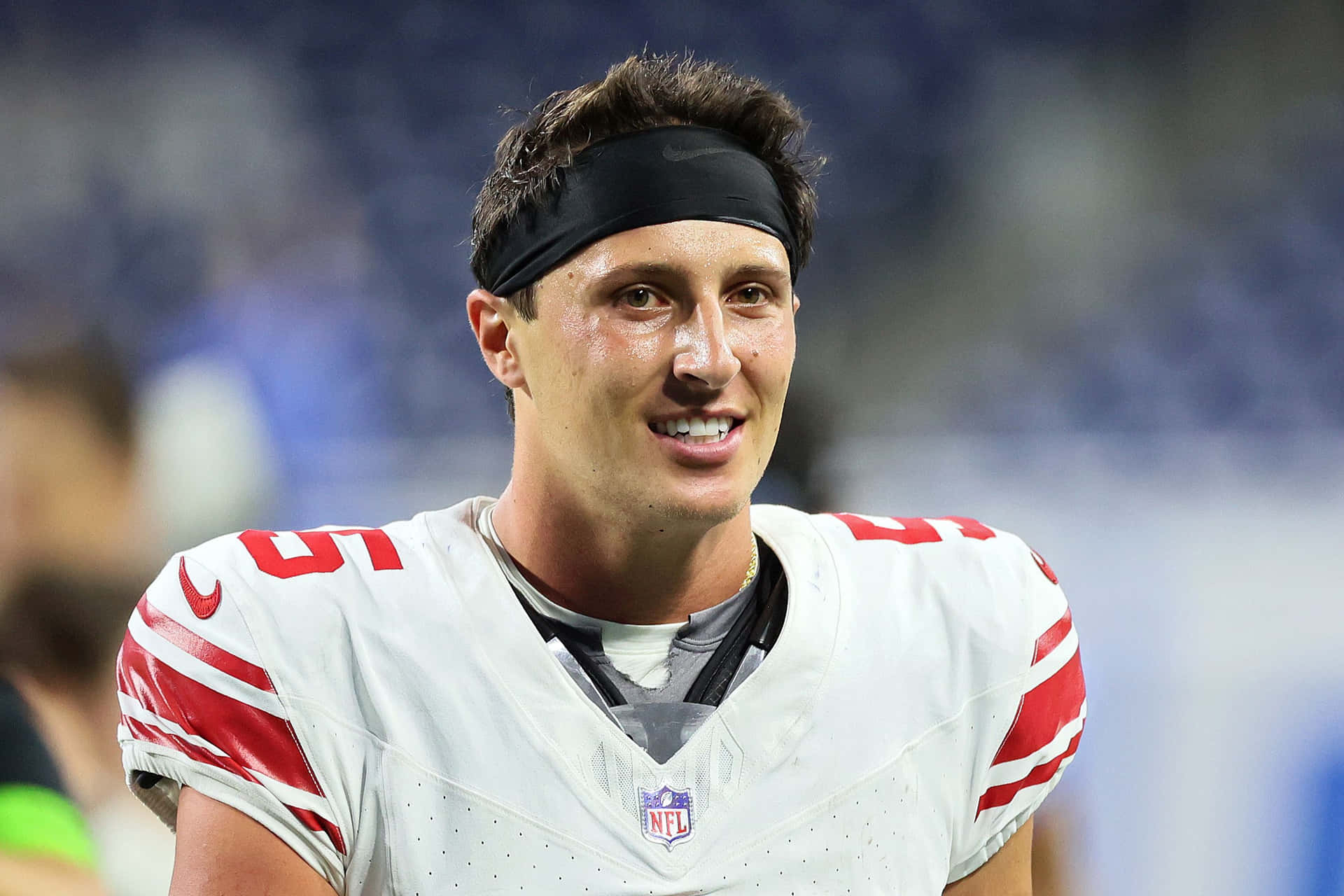  壁紙 — 一位微笑的NFL美式足球員，身穿白色和紅色97號球衣，戴著黑色頭帶，在體育場球場上。
