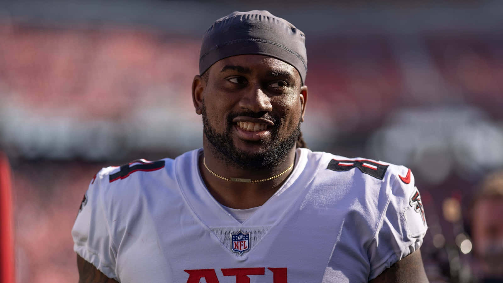  壁紙 — 一位微笑的NFL美式足球員，身穿白色亞特蘭大獵鷹隊球衣，紅色字樣和灰色頭帶，站在陽光明媚的球場上。