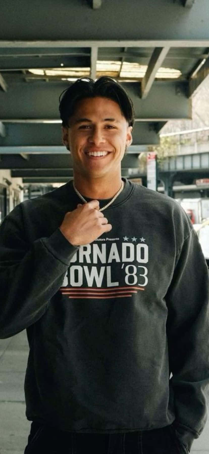  壁紙 — 一位微笑的年輕男子穿著印有「TORNADO BOWL '83」字樣的深色運動衫，站在城市天橋下。