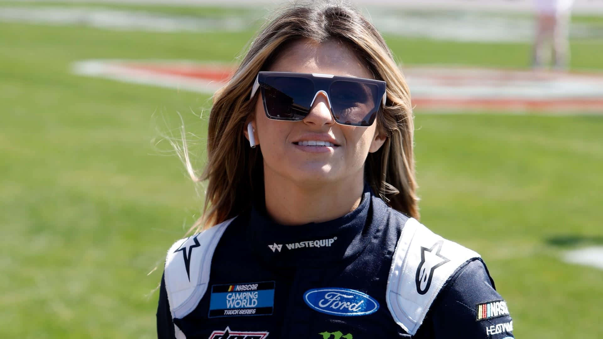  壁紙 — 一位微笑的女NASCAR車手穿著福特賽車服和太陽眼鏡，自信地站在草地賽道旁。