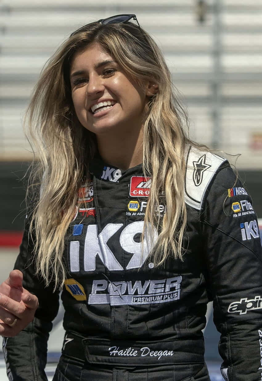  壁紙 — 一位微笑的女性NASCAR車手，身穿黑色賽車服，上面有贊助商標誌，自信地站在賽道上。